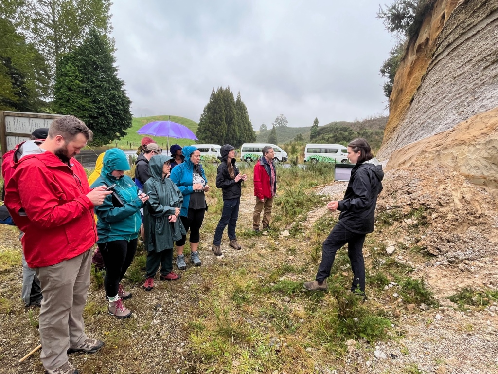 Ellen Datlow stands in front of a yellow and white tephra outcrop, instructing the group, who holding phones or tablets. Everyone is dressed in rain gear. Three white vans with University of Waikato branding are visible in the background.